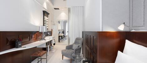 A hotel room with a bed, a vanity with a mirror, a chair, a small table, and a desk with a lamp.