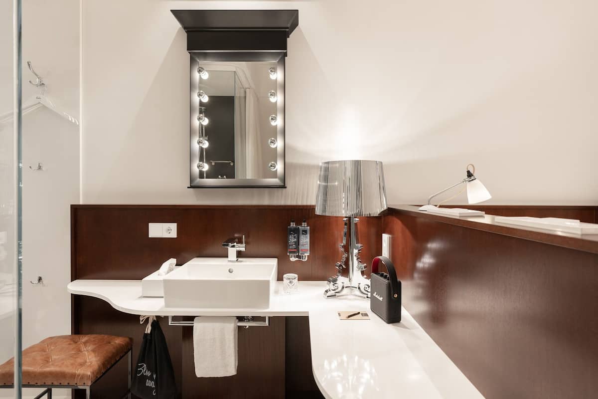 Un baño con tocador, lavamanos, espejo con iluminación, perchero para toallas y un banco.