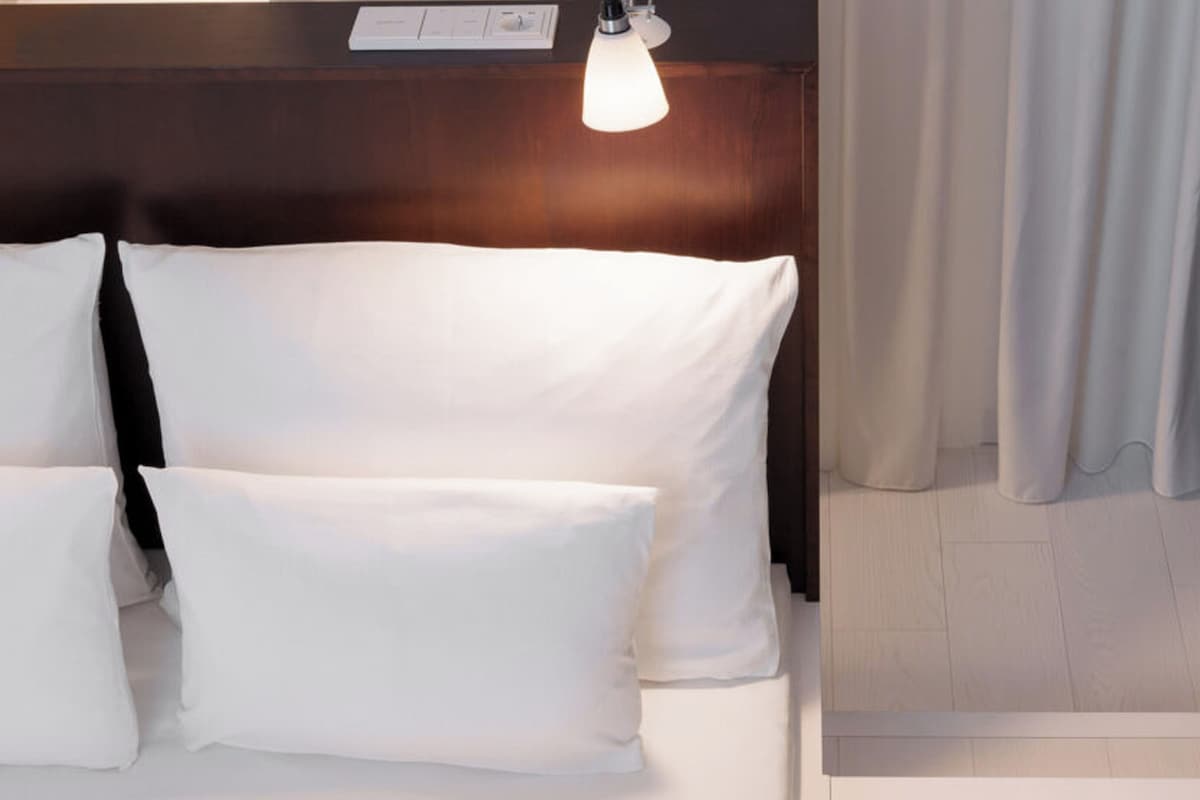 Une chambre d’hôtel avec un lit, des oreillers, une table de chevet, une lampe et un rideau.