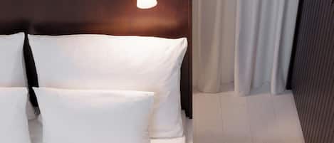 A hotel room with a bed, pillows, a nightstand, a lamp, and a curtain.