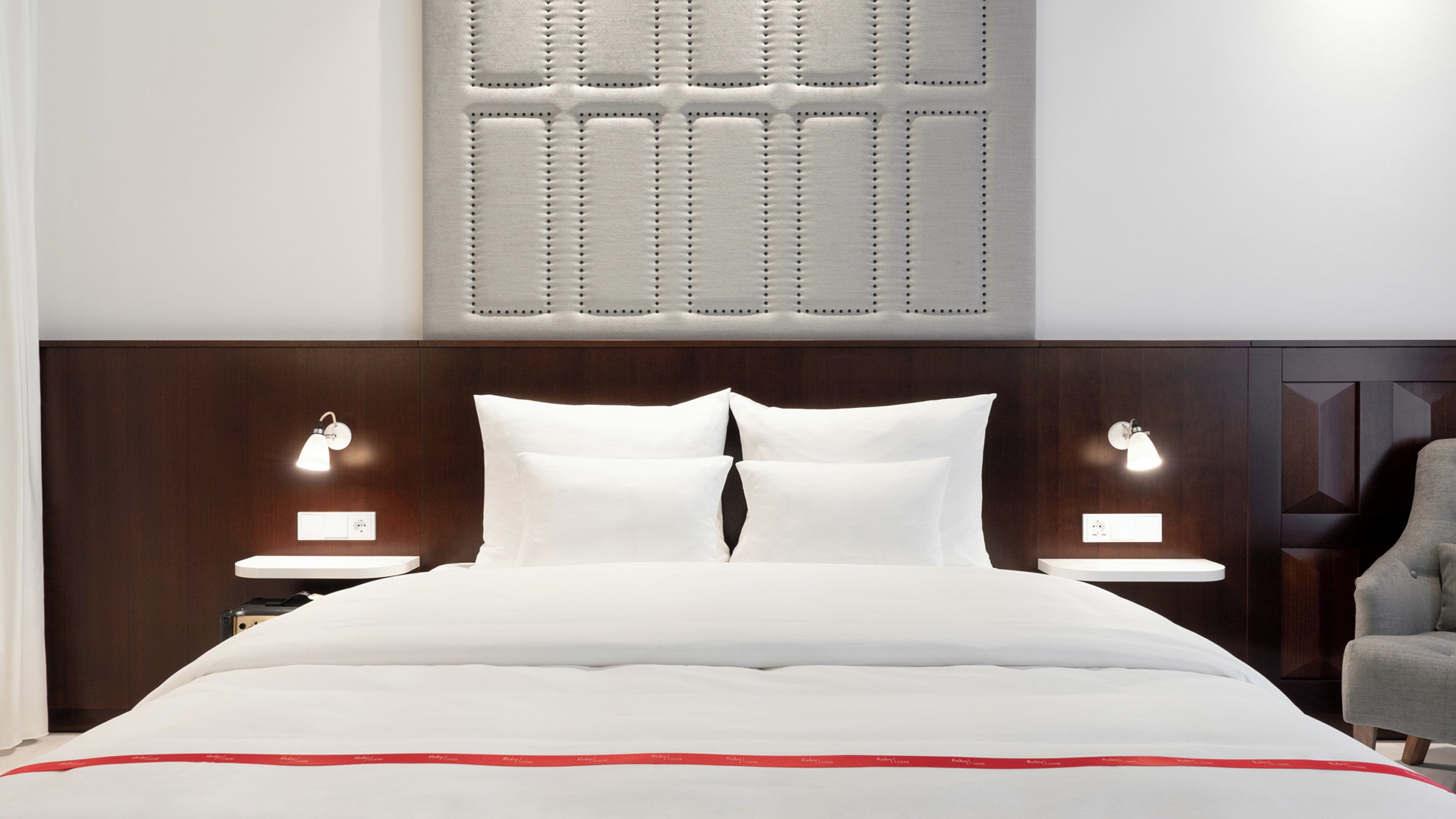 A hotel room with a large bed, two bedside tables with lamps, a wooden headboard, and a grey armchair.