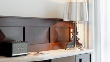 Ein Hotelzimmer mit weißem Schreibtisch, einem schwarzen Marshall-Lautsprecher, einer Lampe mit geriffeltem Schirm und einem hölzernen Kopfteil.