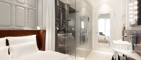 Una habitación de hotel moderna con una cama grande, un vestidor amplio con paredes de cristal, un baño con tocador y un espejo grande.