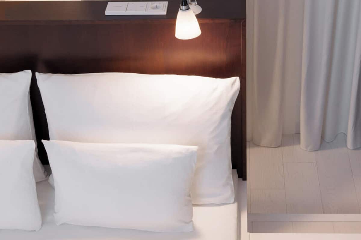 Una habitación de hotel con cama, almohadas, mesita de noche, lámpara y cortina.