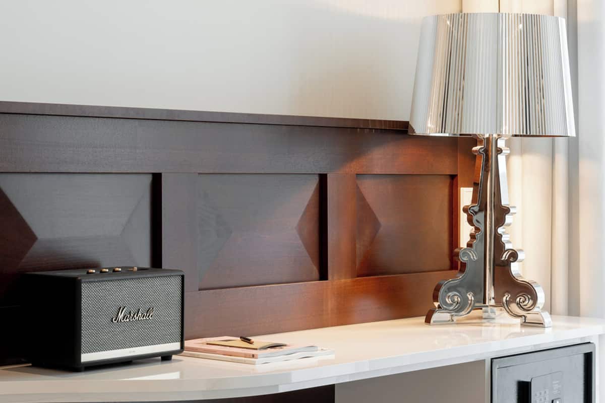 Habitación de hotel con un escritorio blanco, un altavoz Marshall negro, una lámpara con pantalla texturizada y un cabecero de madera.