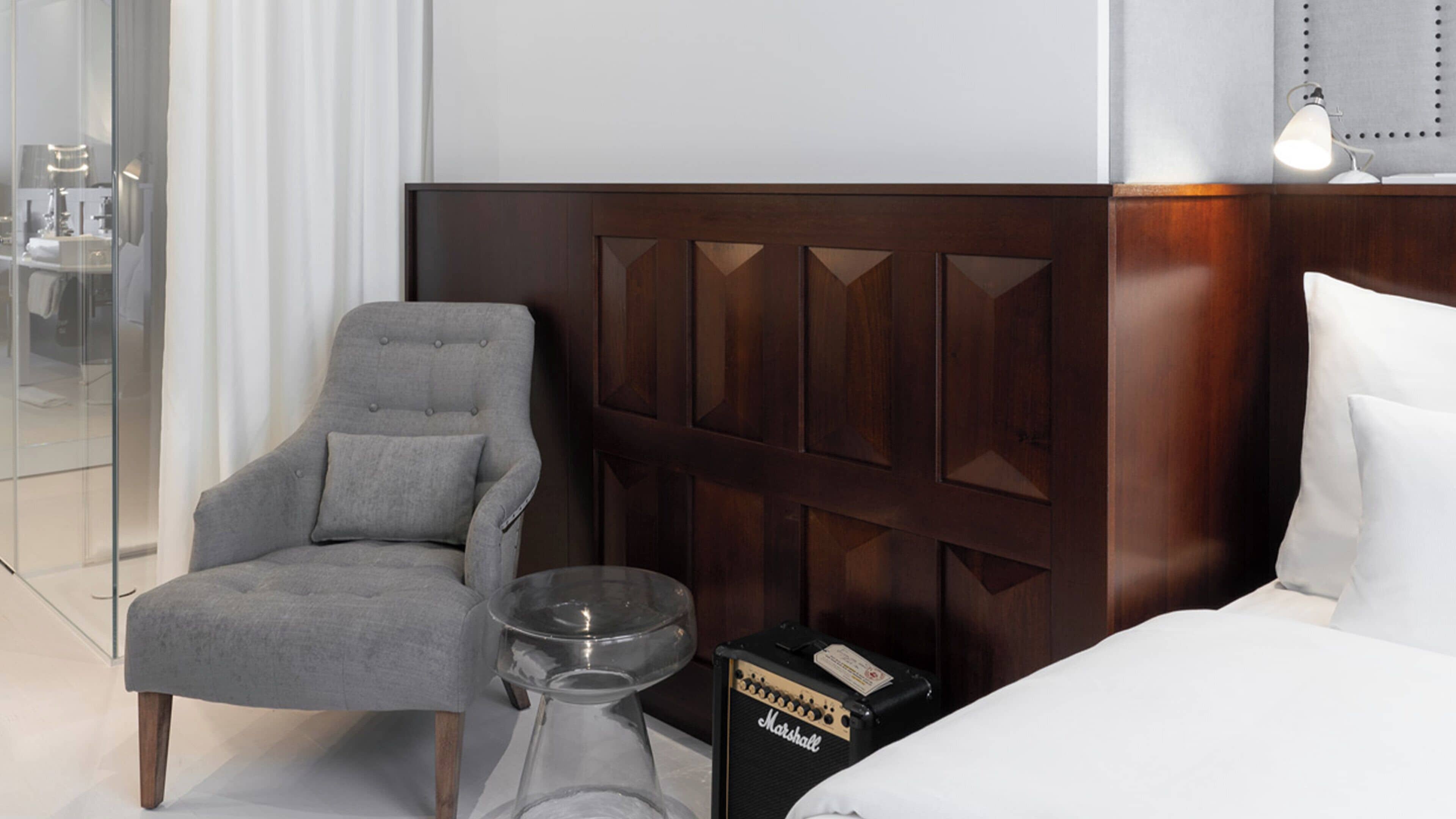 A hotel room with a grey armchair, a bedside table with a glass decanter, a Marshall amplifier, and a bed with white bedding.