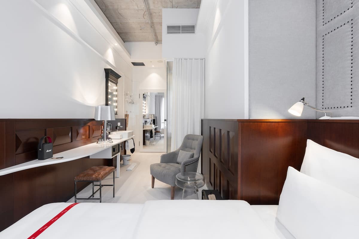 Una habitación de hotel con cama, escritorio con lámpara, silla y tocador con espejos.