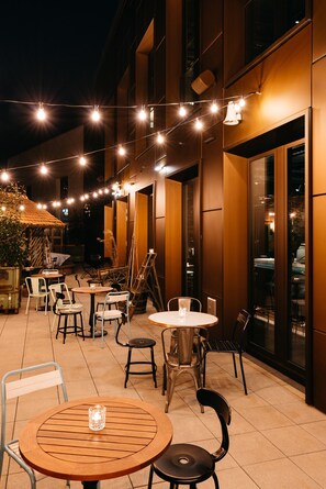 Un espace extérieur aménagé en terrasse, avec des tables et des chaises, des guirlandes lumineuses et un bâtiment doté de grandes fenêtres.