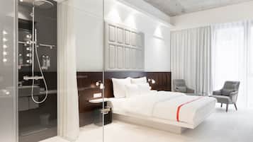 Une chambre d’hôtel moderne dotée d’une douche en verre, d’un grand lit, d’une coiffeuse avec éclairage, d’une tête de lit avec espace de rangement et de deux fauteuils.