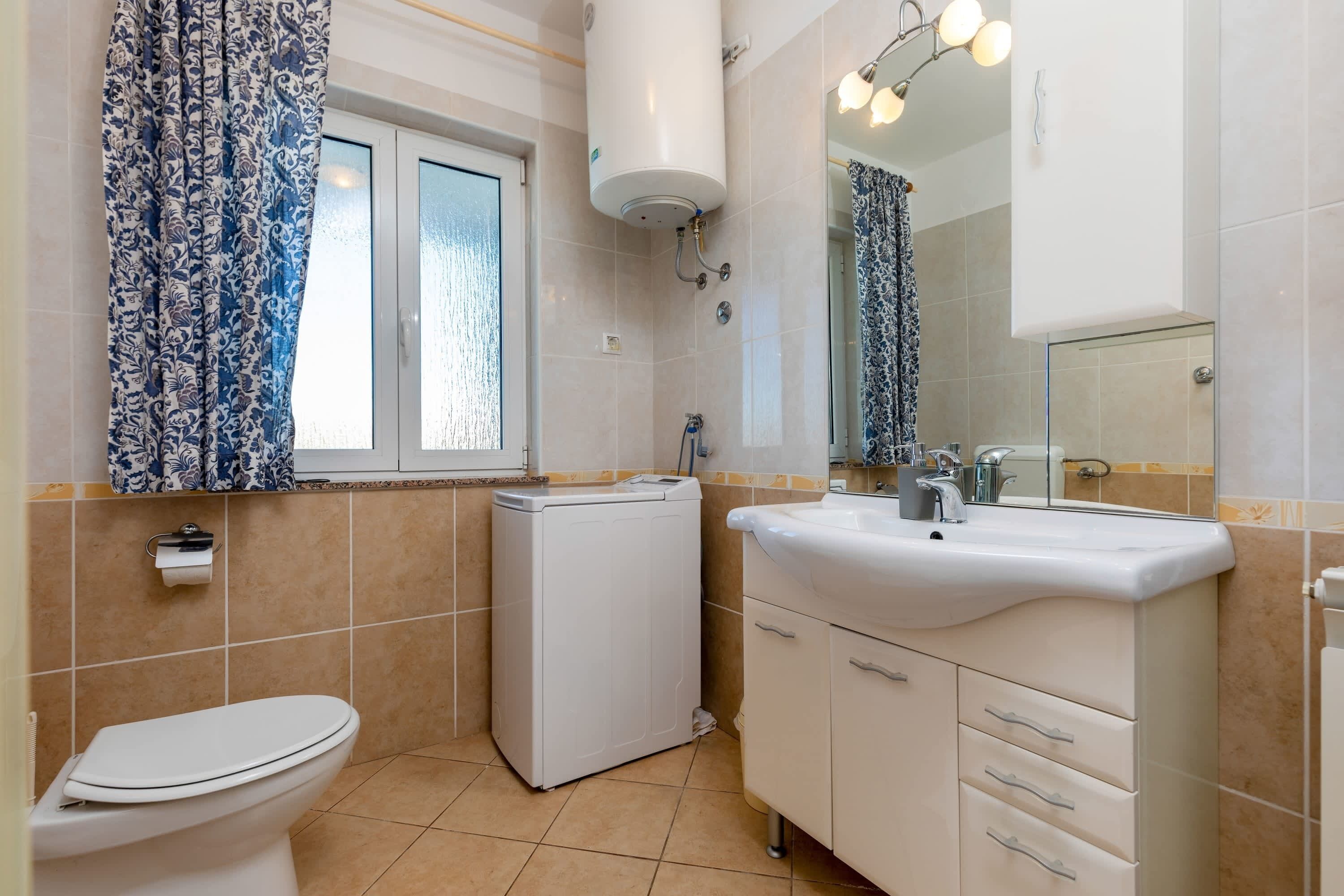 A bathroom with a toilet, washing machine, sink, and a mirror with lights above.