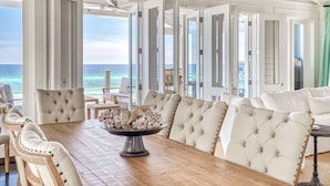 A spacious room with a large wooden dining table, white upholstered chairs, and a view of the ocean through open doors. - Gulf Front in the Middle of Seaside! Elevator, Community Pools, Bikes, and More (Santa Rosa Beach)