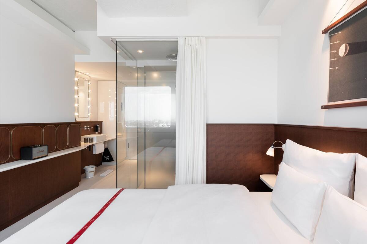 A modern hotel room with a bed, a vanity with a mirror, a shower area, and a view through a glass door.