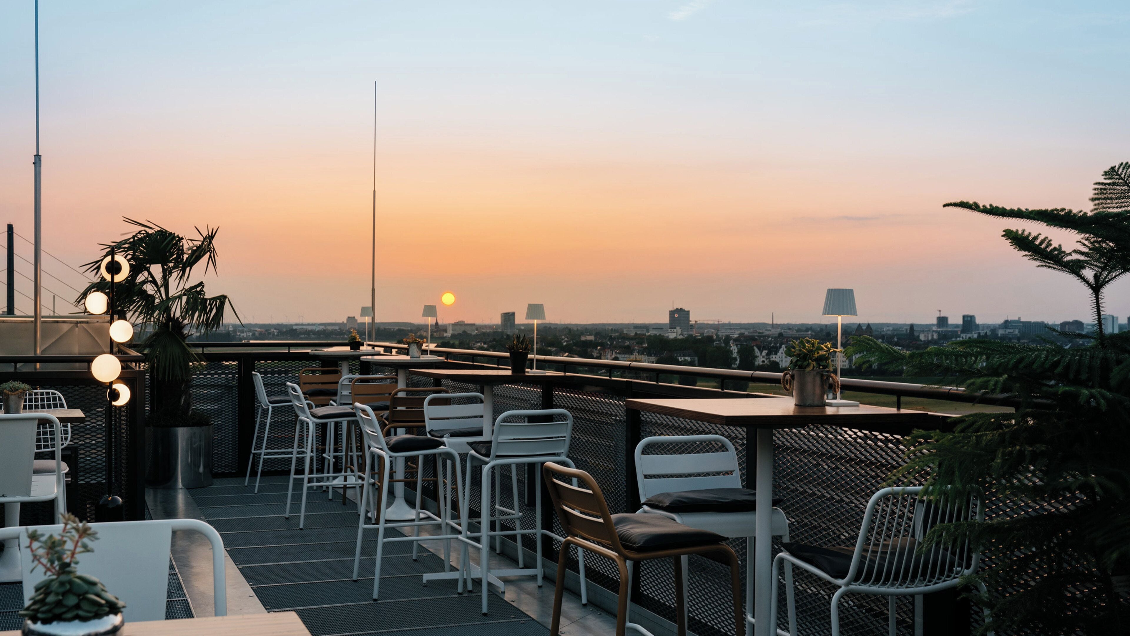 Eine Dachterrasse mit Stühlen und Tischen, Blick auf den Sonnenuntergang und Stadtlandschaft im Hintergrund.