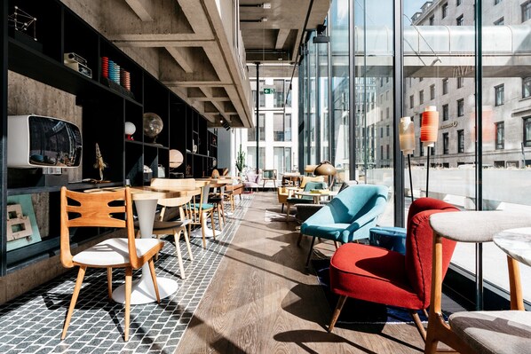 A modern interior with a wooden floor, tables, chairs, and a large window offering a view of the city.