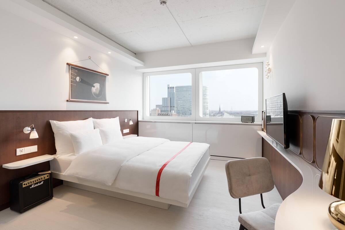 A modern hotel room with a bed, a desk, a chair, a television, and a large window with a city view.