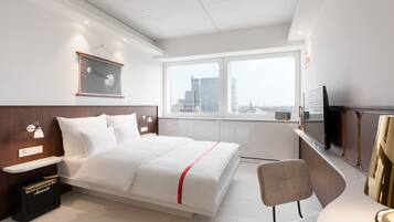 A modern hotel room with a bed, a desk, a chair, a television, and a large window with a city view.