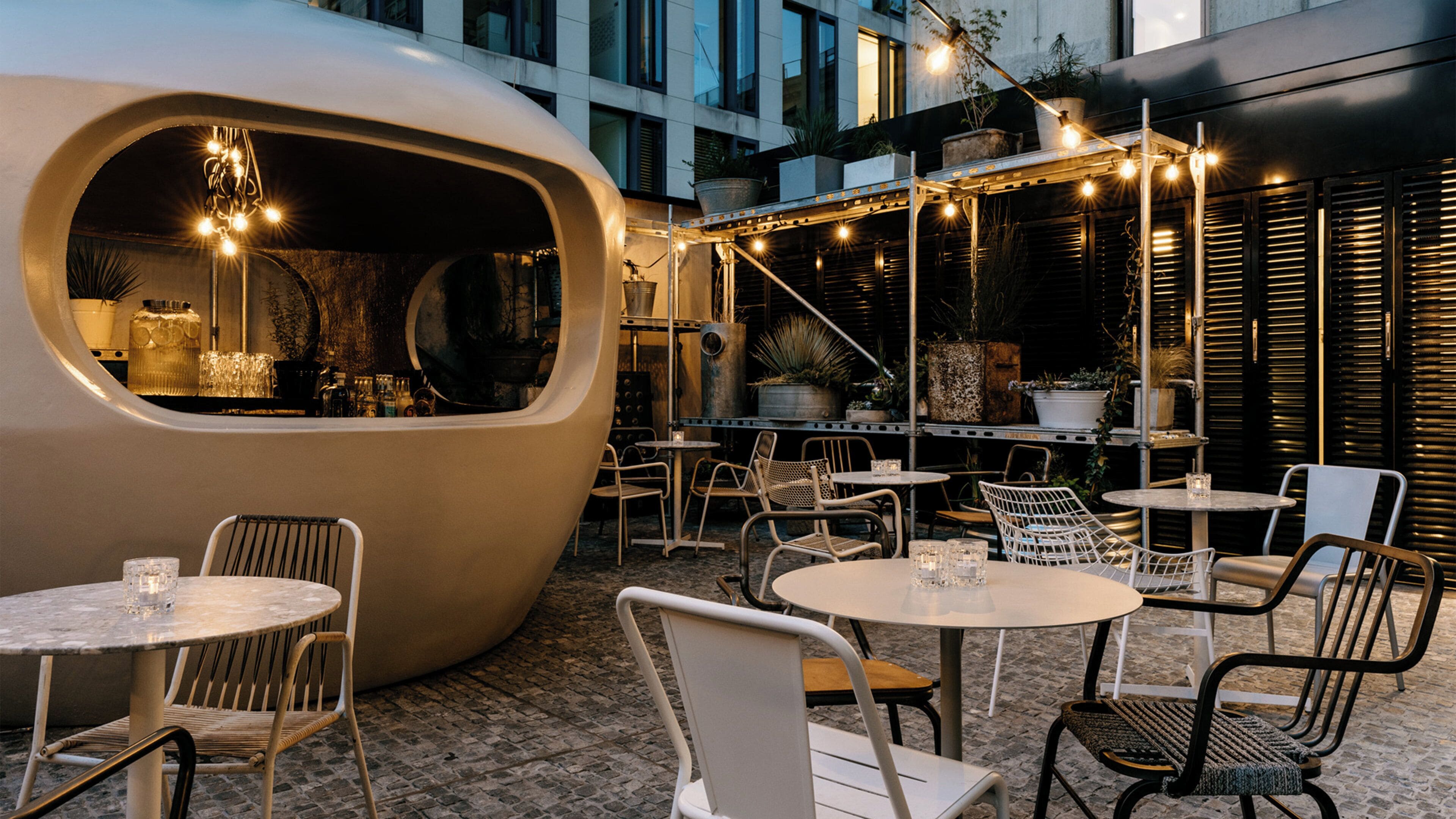 An outdoor seating area with round tables and chairs, a large curved structure with a window, and string lights.