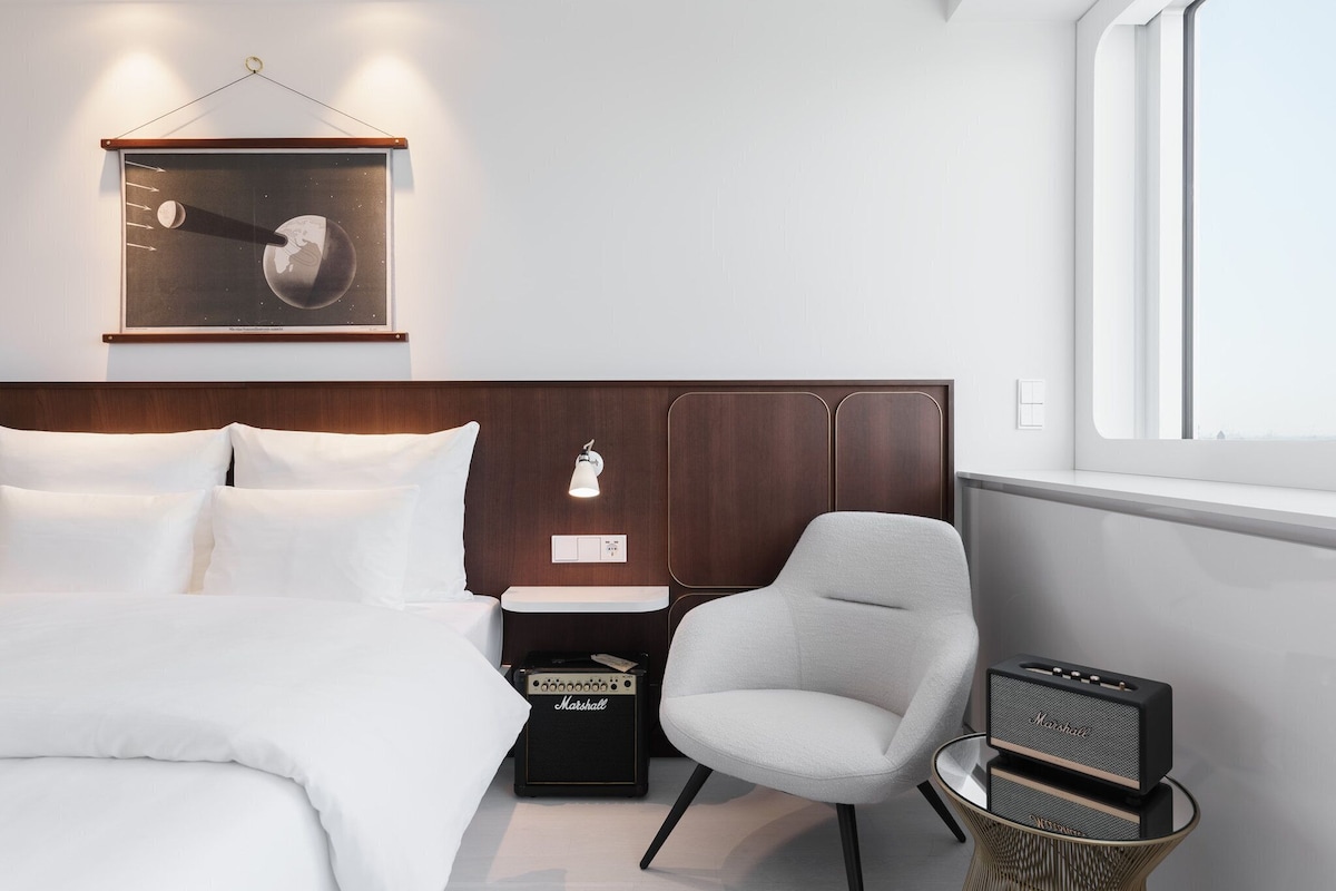 Un dormitorio moderno con cama, una silla, una mesita con un altavoz y un cuadro en la pared.