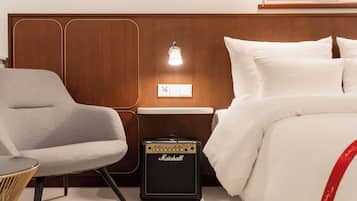 Una camera d'hotel con una testiera in legno, un letto con biancheria bianca, una poltrona grigia e un amplificatore Marshall.