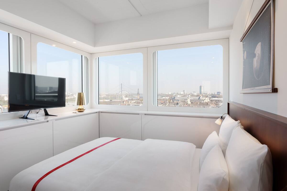 A hotel room with a bed, a TV, and large windows offering a city view.