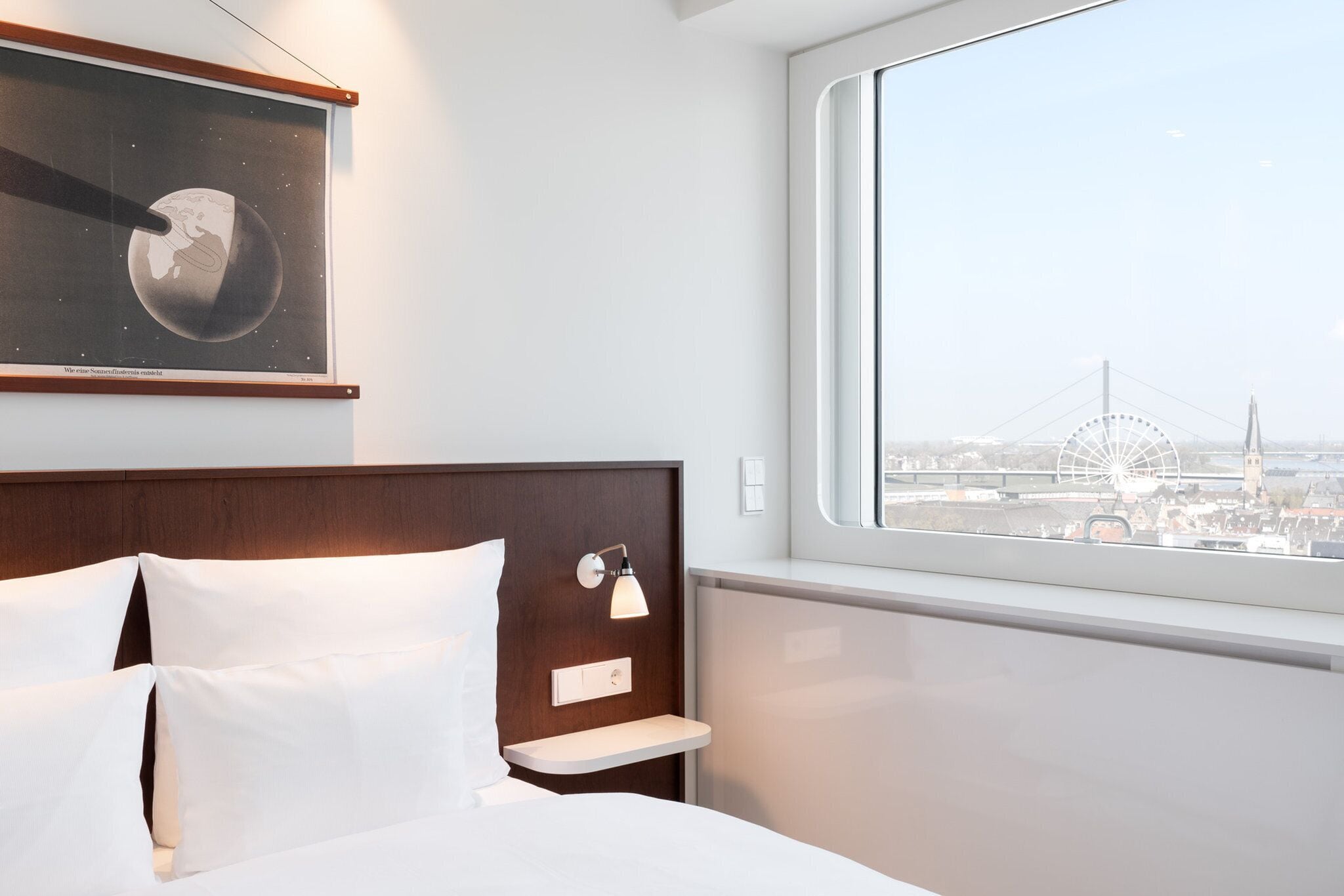 Camera d'albergo con un letto, un comodino, una lampada a parete, una foto incorniciata della Terra e una finestra con vista sulla città.