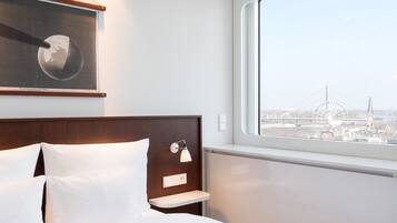 Camera d'albergo con un letto, un comodino, una lampada a parete, una foto incorniciata della Terra e una finestra con vista sulla città.