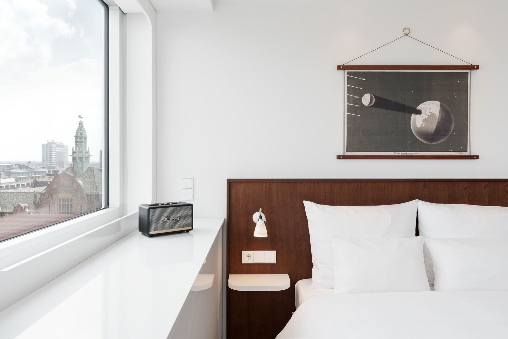 Une chambre d’hôtel moderne dotée d’une grande fenêtre, d’un lit avec du linge de lit blanc, d’une tête de lit en bois, d’une table de chevet avec une lampe et d’un tableau représentant une scène céleste.