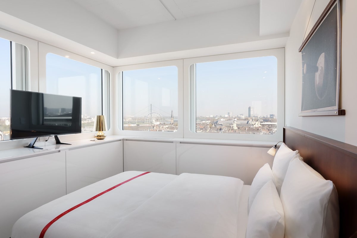 Habitación de hotel con cama, televisión y ventanas amplias que dan a una vista de la ciudad.