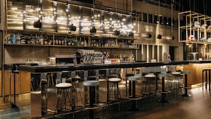 Una zona de bar con taburetes, una barra alargada y diferentes botellas y vasos.