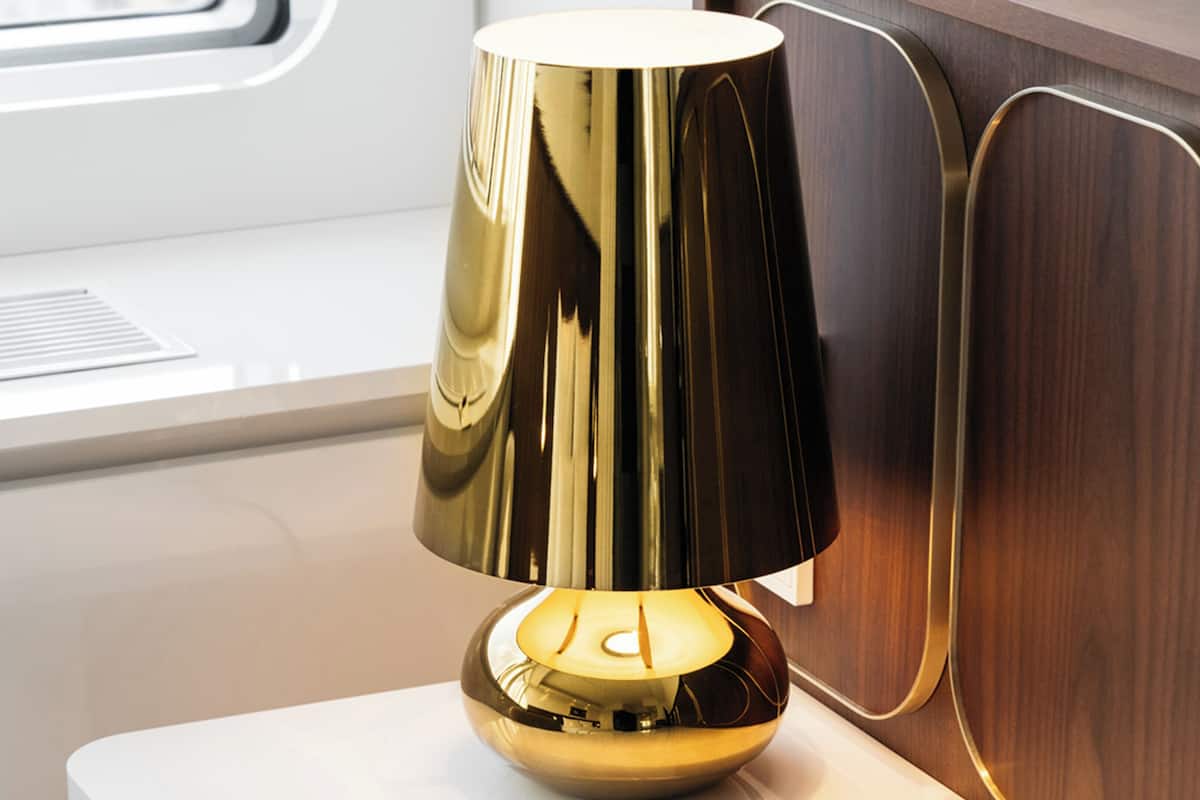 A modern lamp with a reflective gold base and a dark gold lampshade, placed on a white surface near a window with a view of a building.
