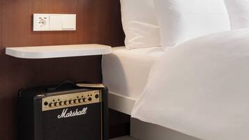 Un lit avec du linge de lit blanc, un amplificateur Marshall, une table de chevet et une prise électrique au mur.