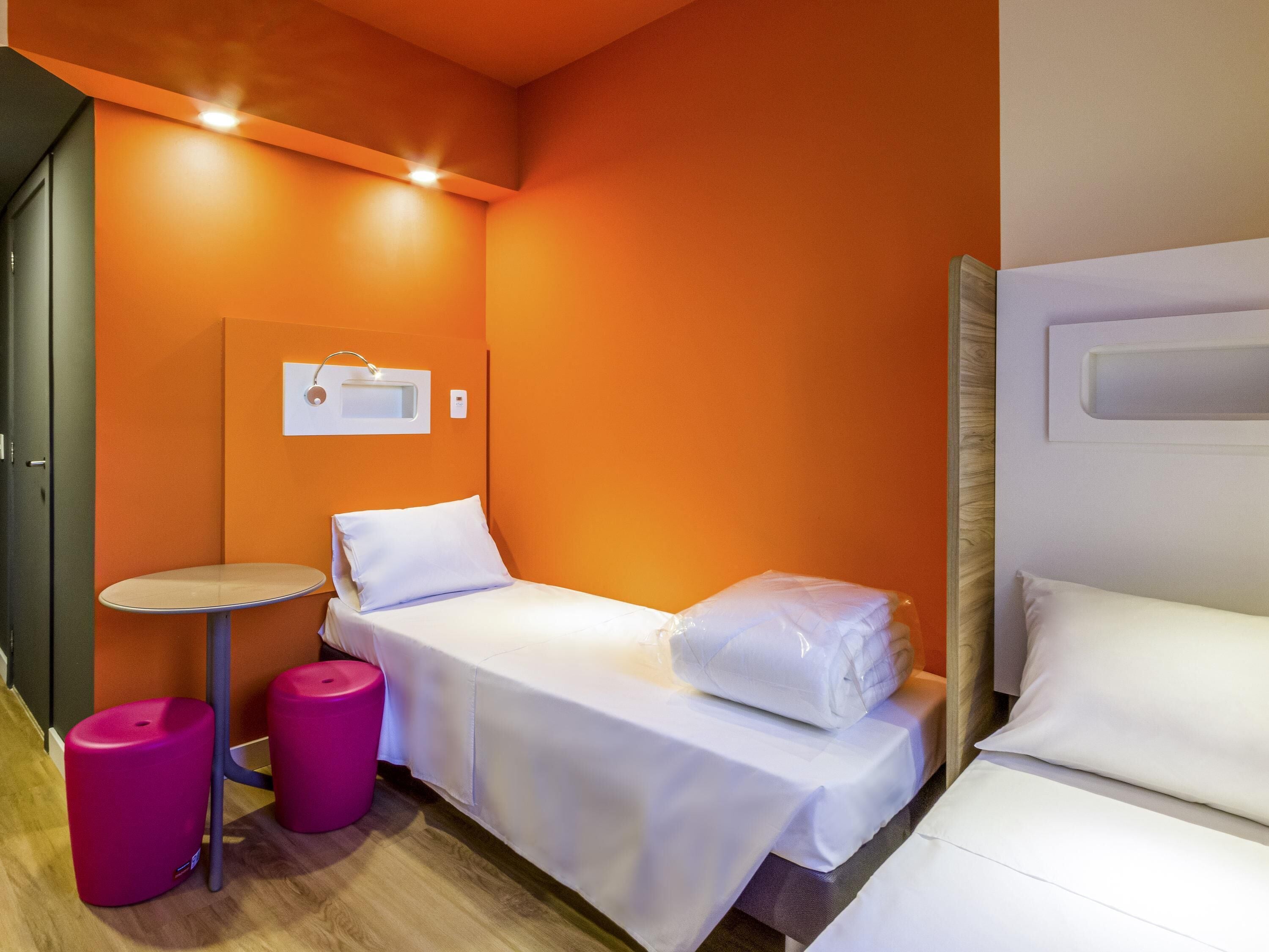 Una habitación pequeña con una cama individual, una pared naranja, una mesita y dos taburetes rosa.