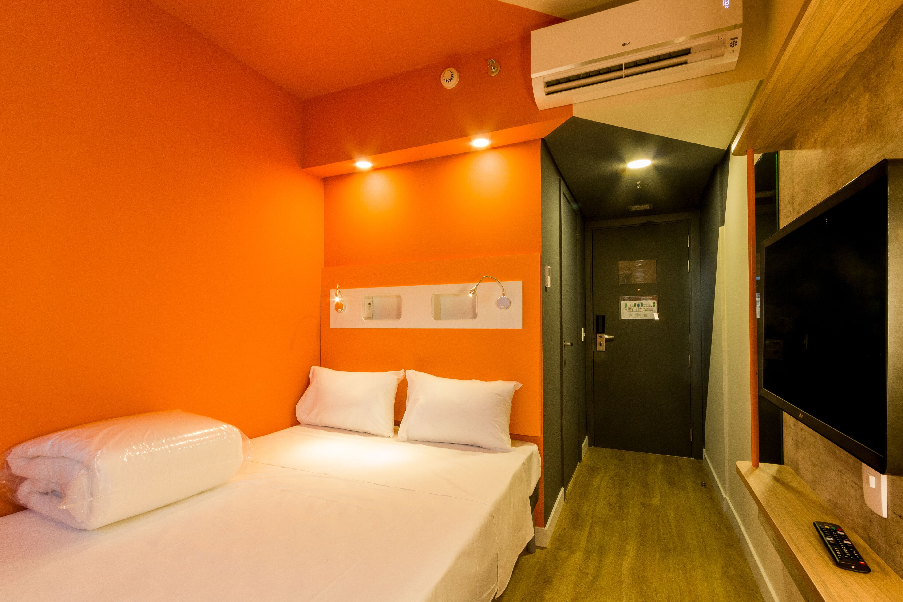 Habitación de hotel con una pared naranja, una cama con ropa de cama blanca, aire acondicionado, televisor y un control remoto sobre una repisa de madera.
