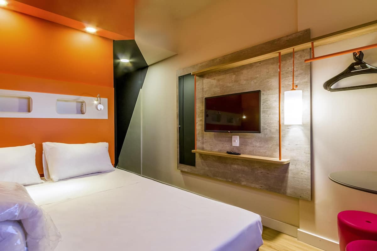 Hotelkamer met een bed, een aan een houten paneel bevestigde televisie, een klein tafeltje en een rode kruk.