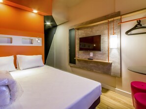 Quarto de hotel com uma cama, uma TV fixada em um painel de madeira, uma mesa pequena e um banquinho vermelho.
