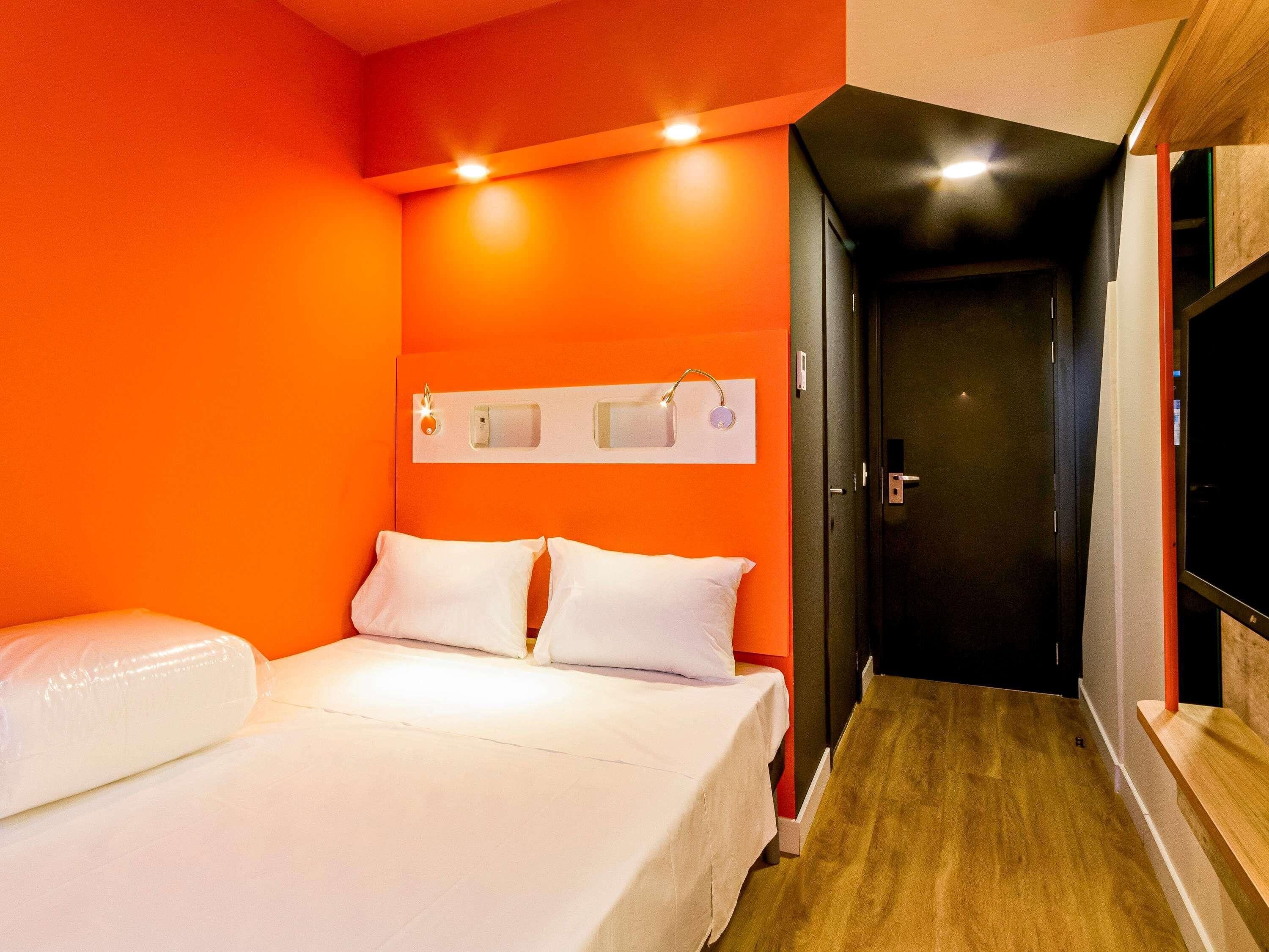 A hotel room with an orange wall, a bed with white bedding, a wooden floor, and a door leading to another room.
