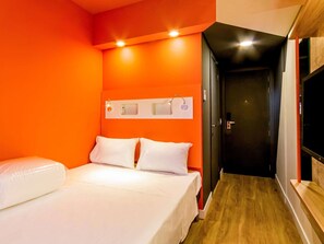 Una habitación de hotel con pared naranja, una cama con ropa de cama blanca, suelo de madera y una puerta que conduce a otra habitación.