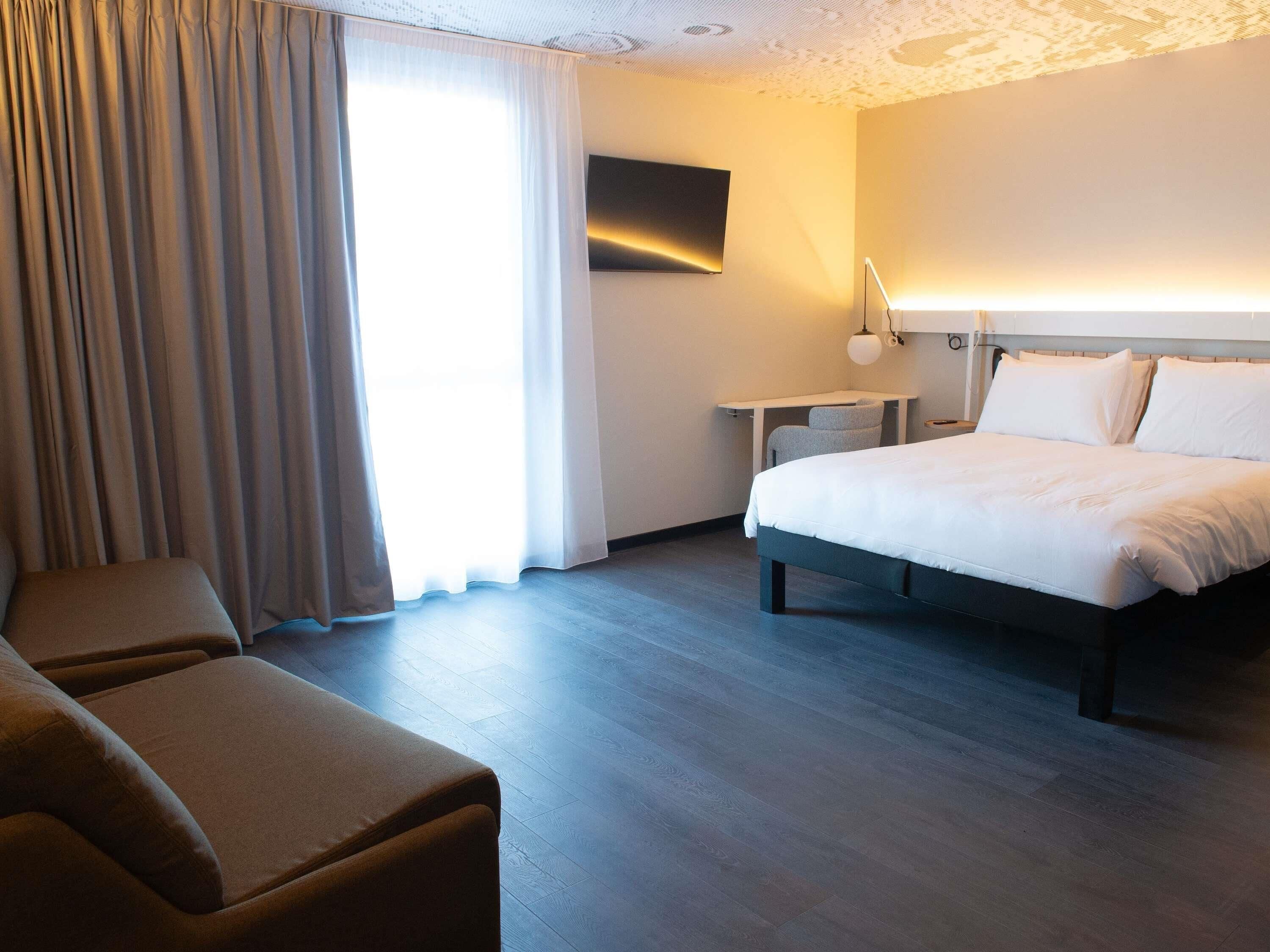 Ein Hotelzimmer mit Bett, Schreibtisch, Stuhl und einem großen Fenster mit Vorhängen.