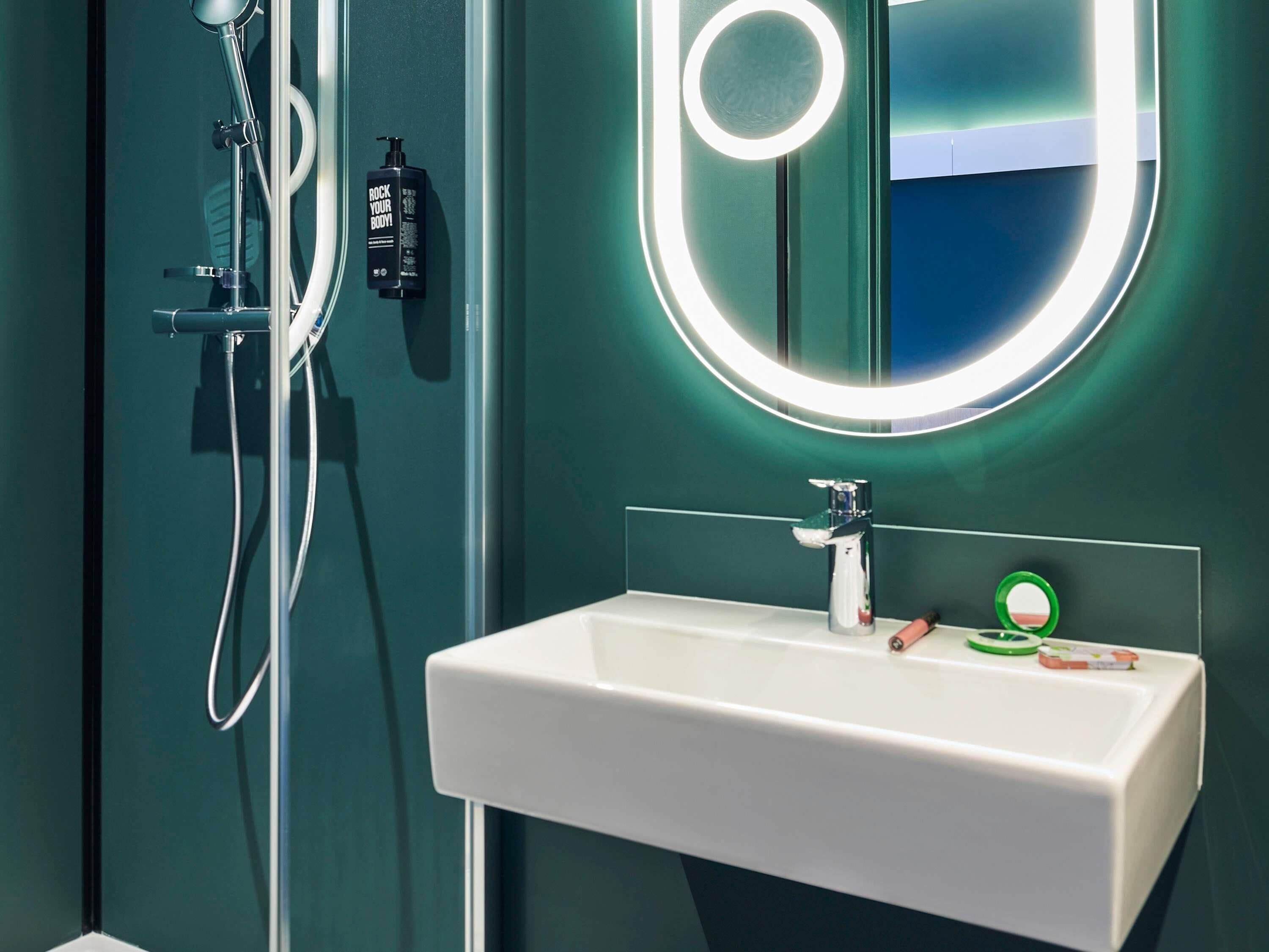 A bathroom with a white sink, a handheld showerhead, a bottle of shampoo, and a mirror with a circular light.