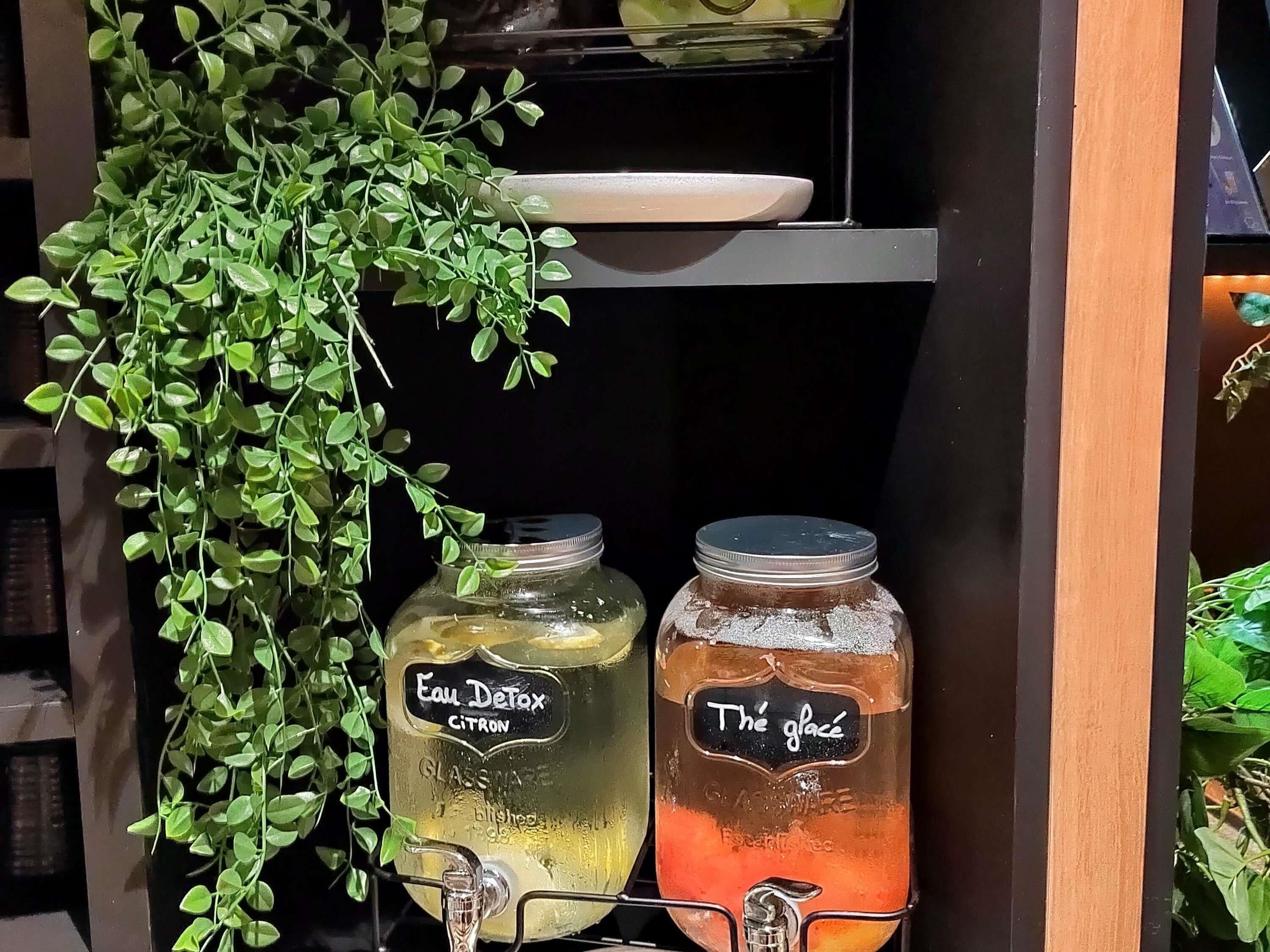 Een plank met twee glazen potten, gelabeld 'Eau Detox CITRON' en 'Thé glace', en een hangende plant.