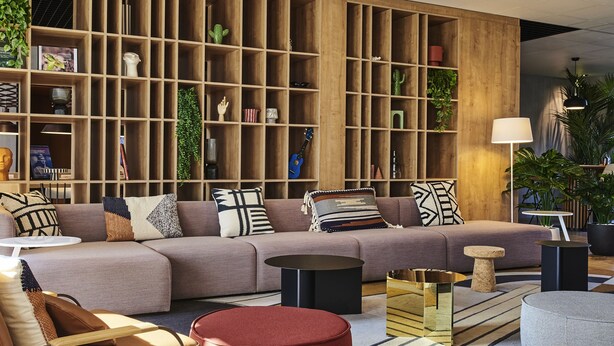 Un salon moderne avec un canapé d’angle, un pouf rouge et une bibliothèque en bois remplie de plantes et de livres.