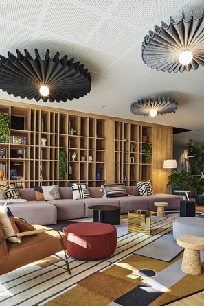 Een moderne woonkamer met een hoekbank, een rode poef en een houten boekenkast vol planten en boeken.