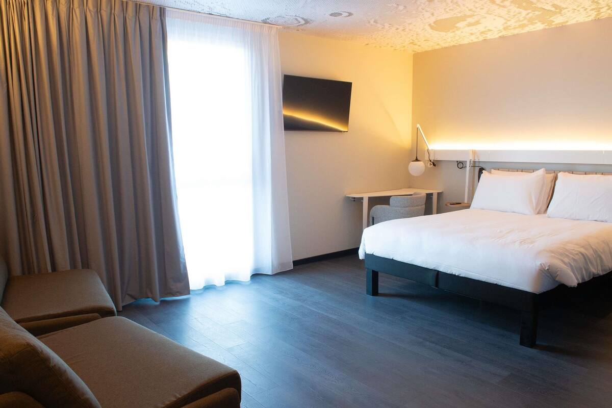 Ein Hotelzimmer mit Bett, Schreibtisch, Stuhl und einem großen Fenster mit Vorhängen.