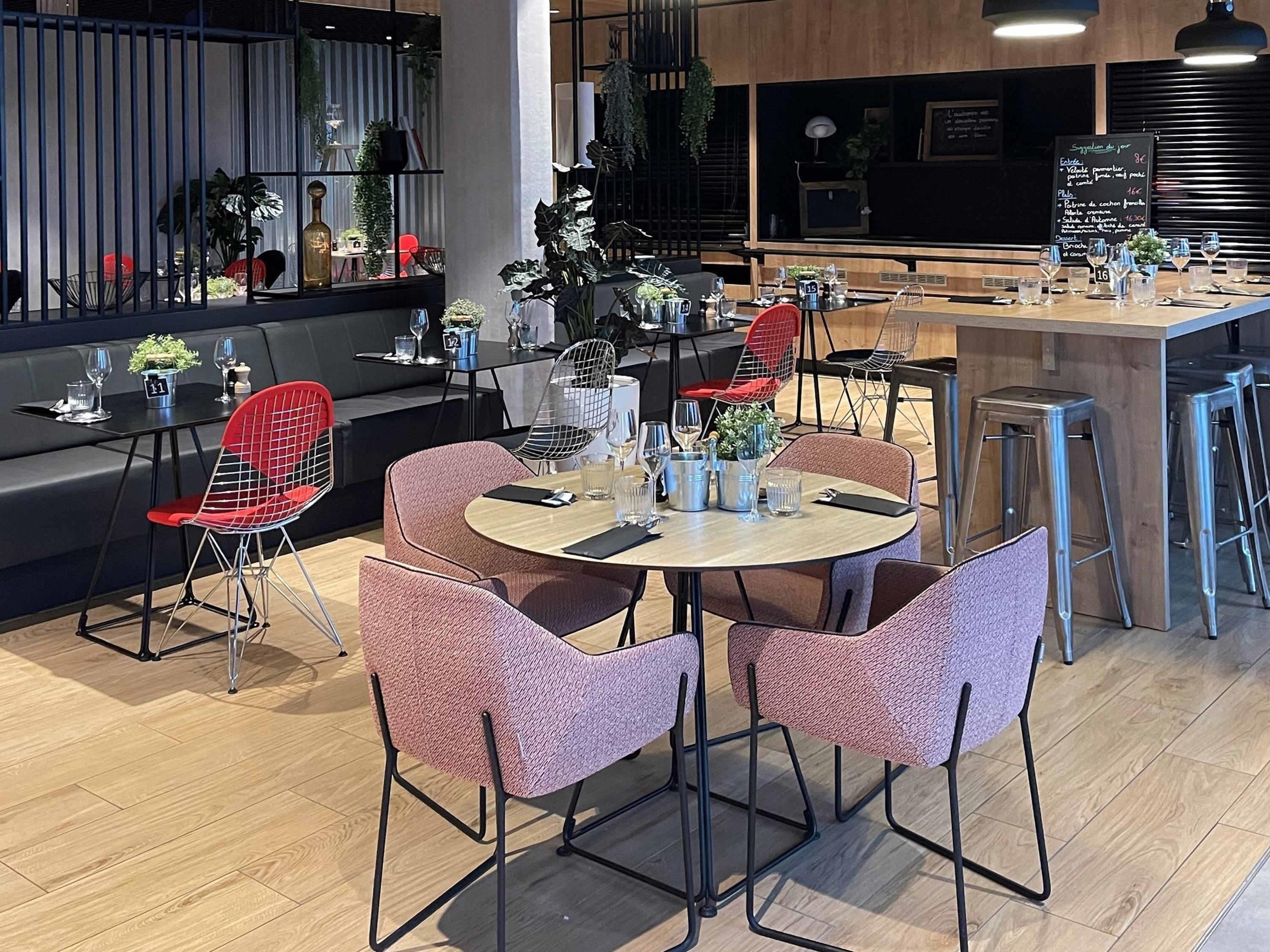 A modern restaurant with round tables, red wireframe chairs, and a bar area.