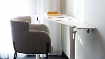 Una habitación moderna con una butaca tapizada en gris, un escritorio blanco con un cuaderno y un libro, y una ventana con cortinas translúcidas.