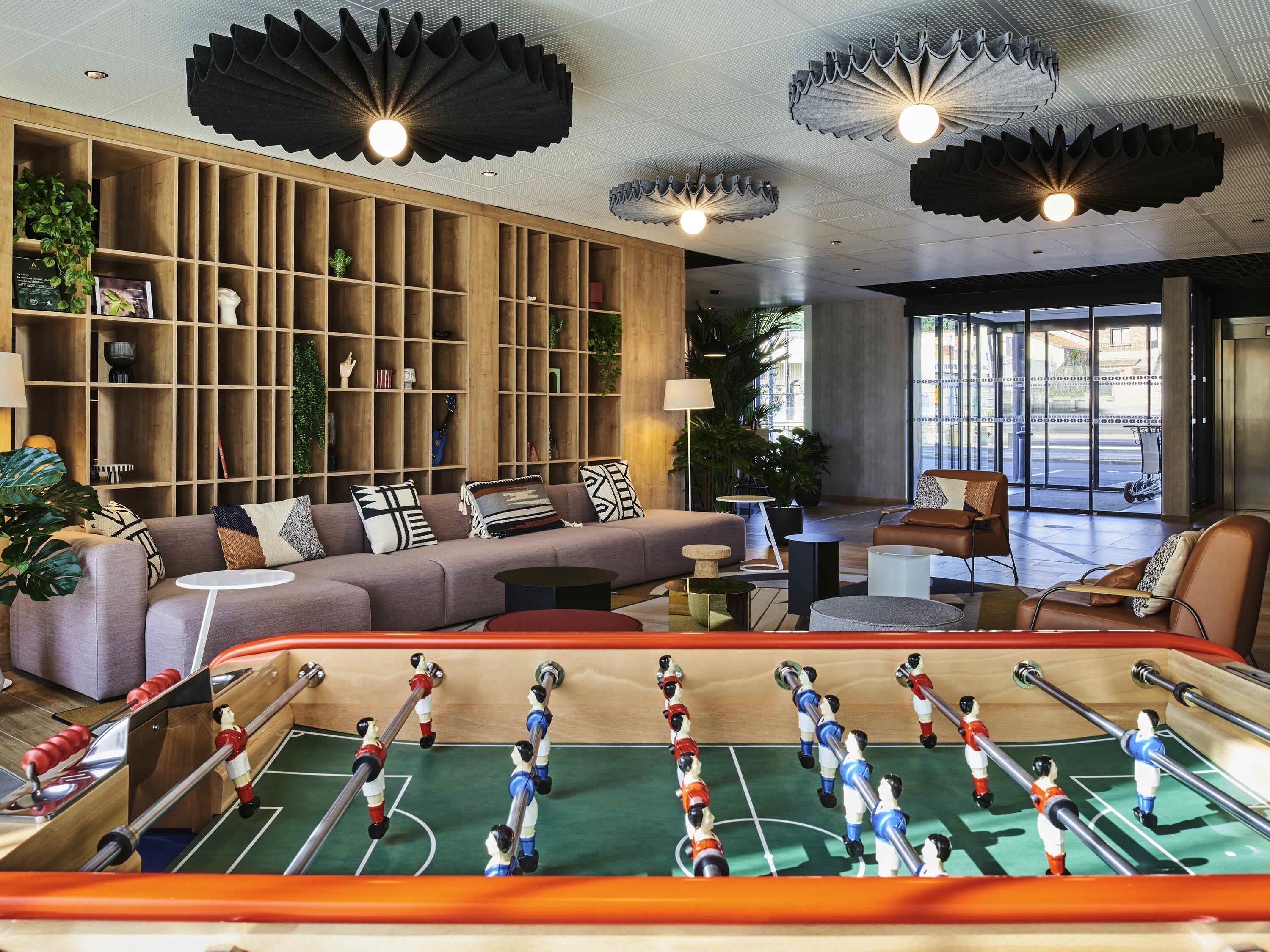 A modern living room with a foosball table, a sofa with patterned cushions, and a wooden shelving unit with various decorative items.