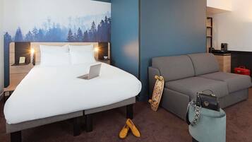 Habitación de hotel con cama, sofá, una laptop sobre la cama, un monopatín, un bolso de mano y una maleta.