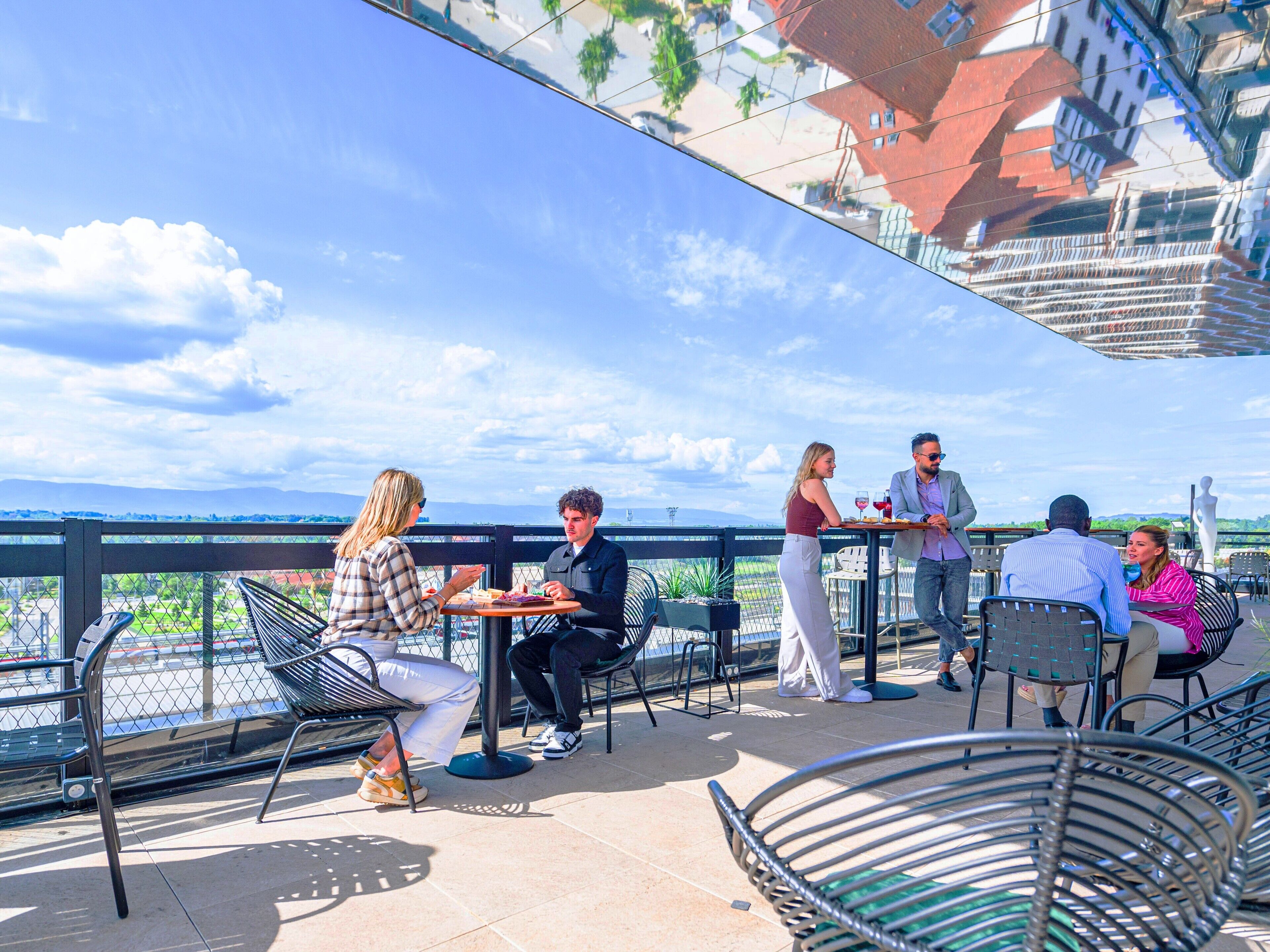 Une terrasse extérieure avec des chaises et des tables en métal, une vue sur le paysage urbain et un ciel dégagé.
