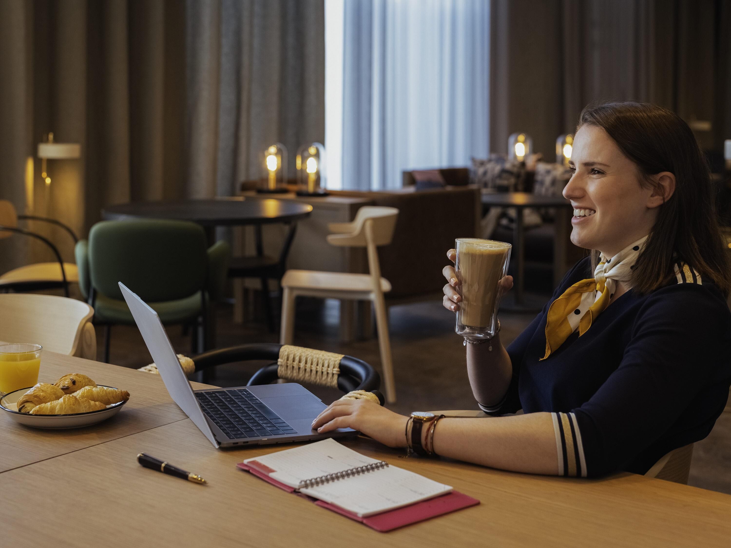A person at a table with a laptop, a notebook, and a glass of coffee. There are chairs and tables in the background, and a window with curtains.