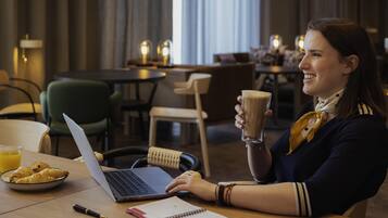 A person at a table with a laptop, a notebook, and a glass of coffee. There are chairs and tables in the background, and a window with curtains.